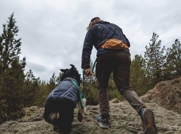 A man wearing the Ruffwear Home Trail Hip Pack and hiking with his dog on a rocky trail with trees in the background