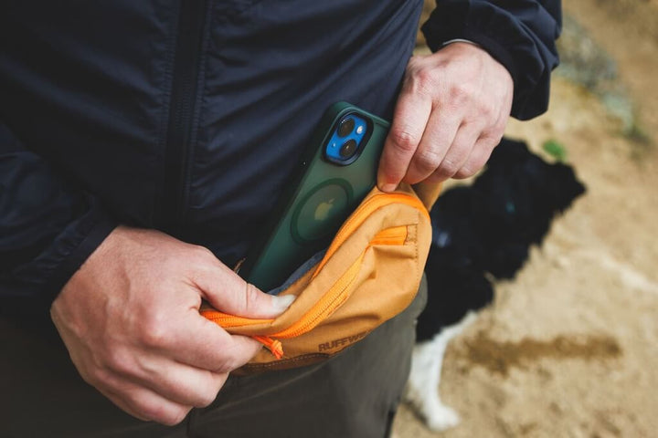 Person placing a phone into a pocket on the Ruffwear Home Trail Hip Pack with a dog in the background